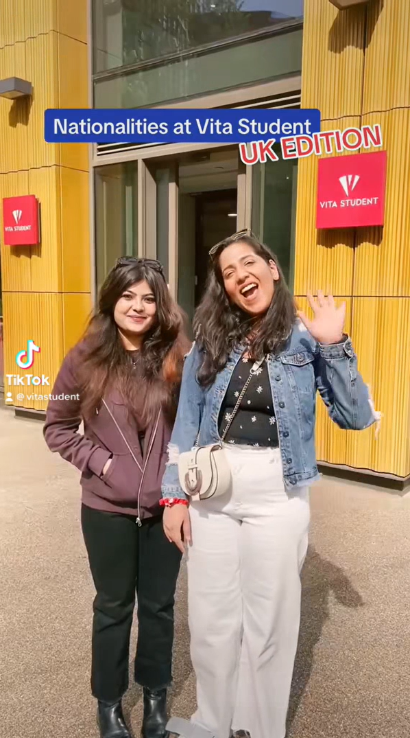 Two people stand in front of a Vita Student building. One wears a dark jacket, the other a denim jacket with white pants, waving and smiling. Banners read "Vita Student." A TikTok logo and overlay say: "Nationalities at Vita Student UK Edition," capturing the vibe of studying abroad in India.